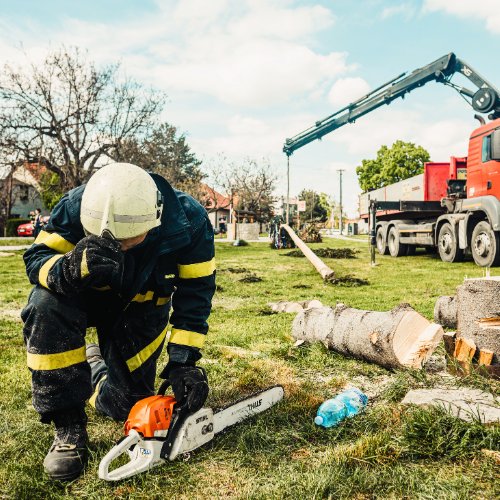 Worker in protective gear kneeling beside a chainsaw, with cut tree sections and a crane truck behind